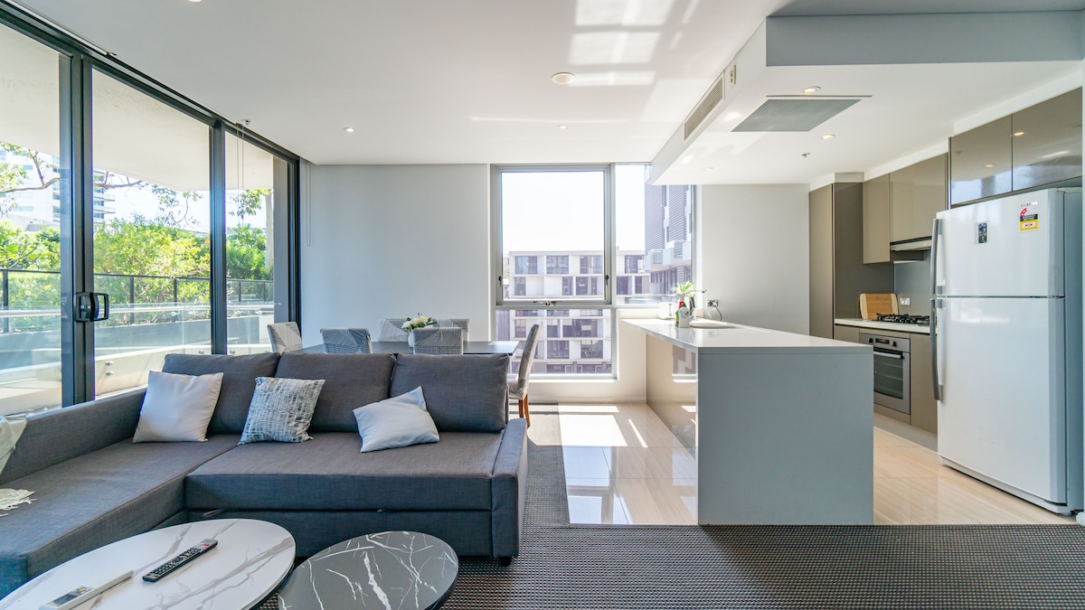 An open-plan living area is showcased, featuring a spacious gray sofa with decorative pillows. A modern kitchen with sleek cabinetry and stainless-steel appliances is visible. Natural light floods the room through large windows, illuminating the polished floors and dining area.