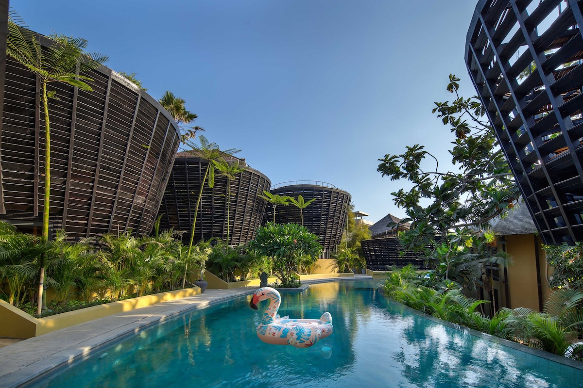 An outdoor swimming pool is surrounded by lush greenery and modern architectural structures. A large inflatable swan floats on the water's surface, reflecting the clear blue sky above. The pool area is designed for relaxation, with palm trees and tropical plants enhancing the serene setting.