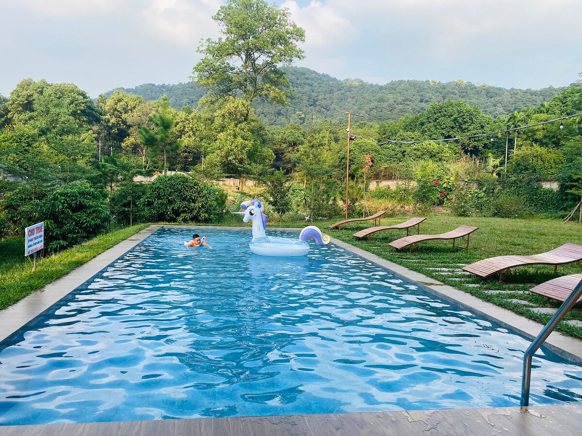 A swimming pool is surrounded by lush greenery, with rolling hills in the background. A unicorn-shaped float is positioned in the water. Nearby, several lounge chairs are arranged for relaxation, creating a welcoming outdoor space for enjoyment.