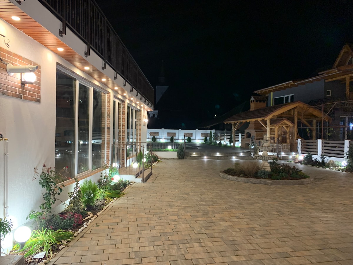 A well-lit outdoor area features a paved walkway, surrounded by neatly arranged landscaping. Soft illumination highlights various plants and decorative elements. The building's large windows reflect the serene ambiance, while nearby structures provide a sense of cozy architecture under the night sky.