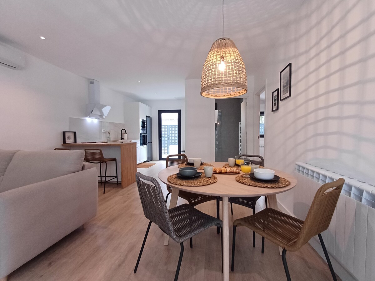 A dining area is presented with a round table surrounded by four chairs. A woven pendant light hangs above, casting soft shadows. A sleek kitchen is visible in the background, featuring modern appliances and an island with bar stools.