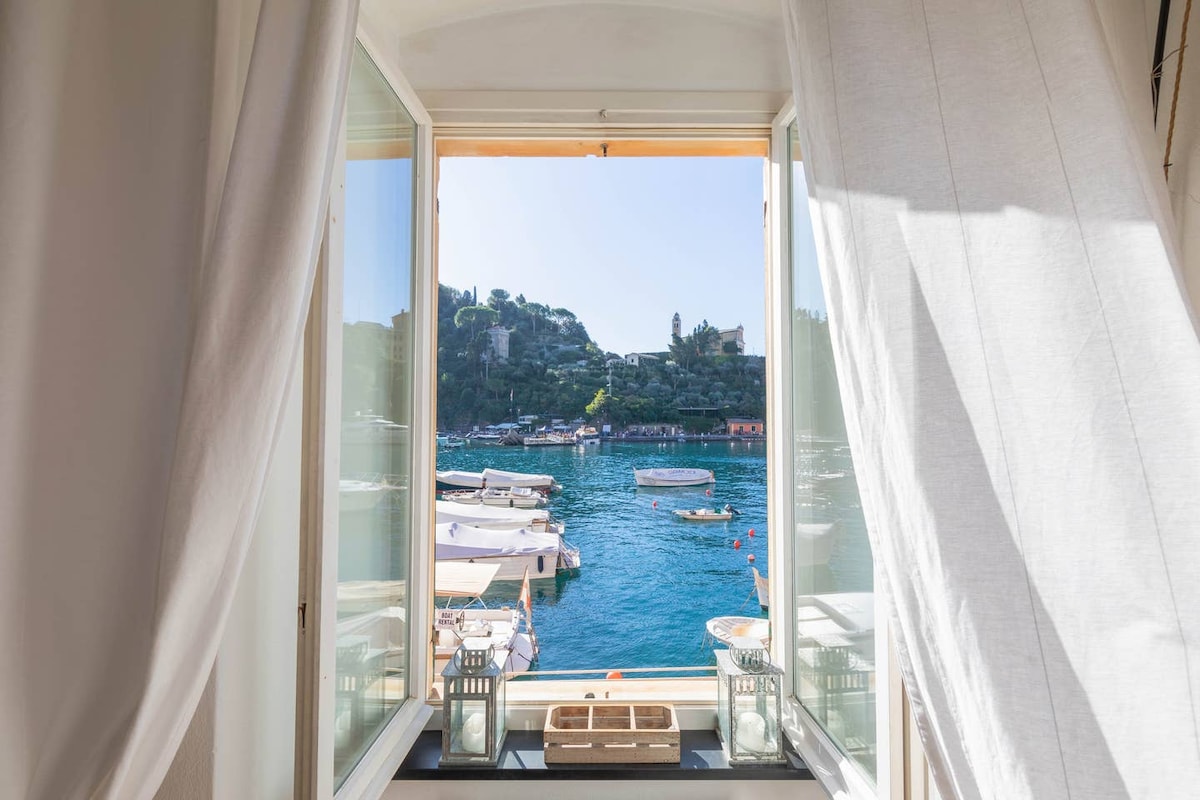 A view from large open windows reveals a vibrant bay with boats gently bobbing on the water. Soft white curtains frame the scene, enhancing the feeling of light and openness.
