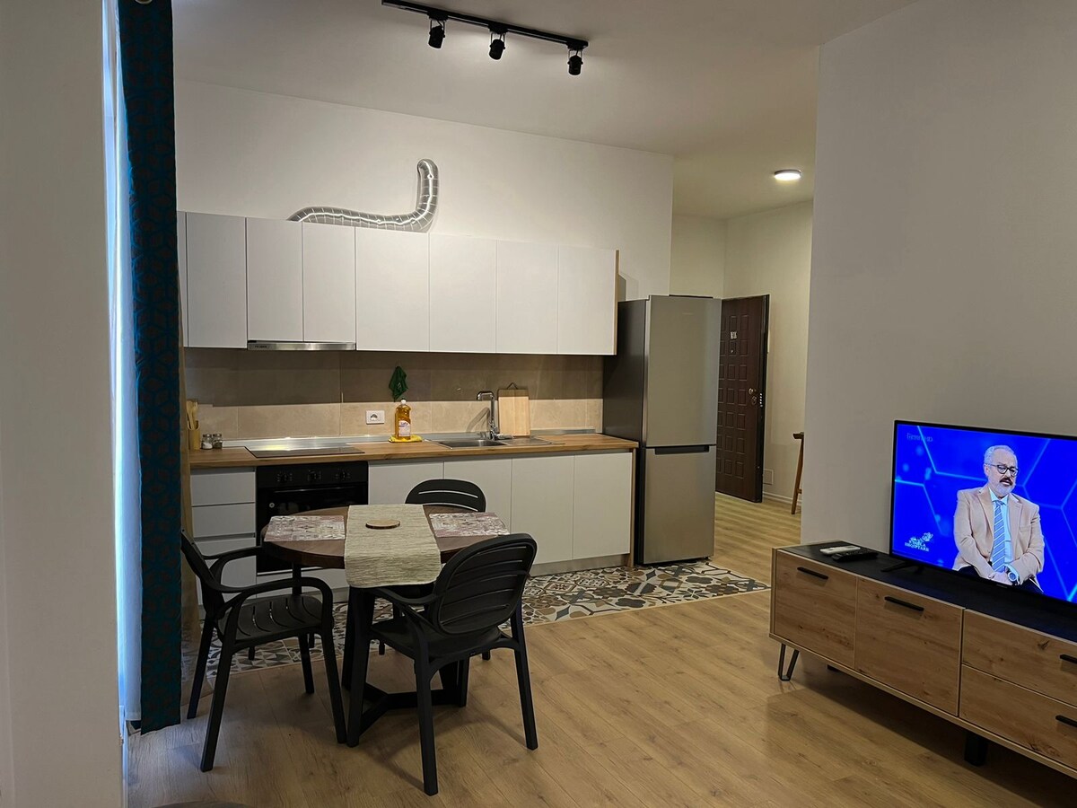 A modern kitchenette is featured with sleek cabinetry and essential appliances. A dining table with chairs is positioned nearby. Light-colored flooring complements the space, while a television screen displays a program in the living area, enhancing the casual atmosphere.