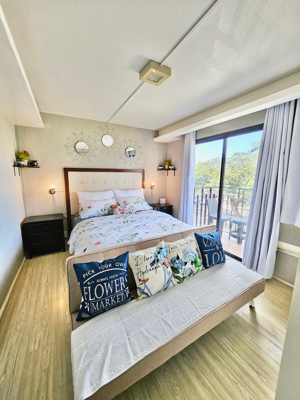 A bright bedroom is shown, featuring a spacious bed dressed in floral linens. A decorative bench is positioned at the foot of the bed, adorned with cushions. Natural light enters through a sliding glass door leading to a balcony, complemented by mirrors on the wall.