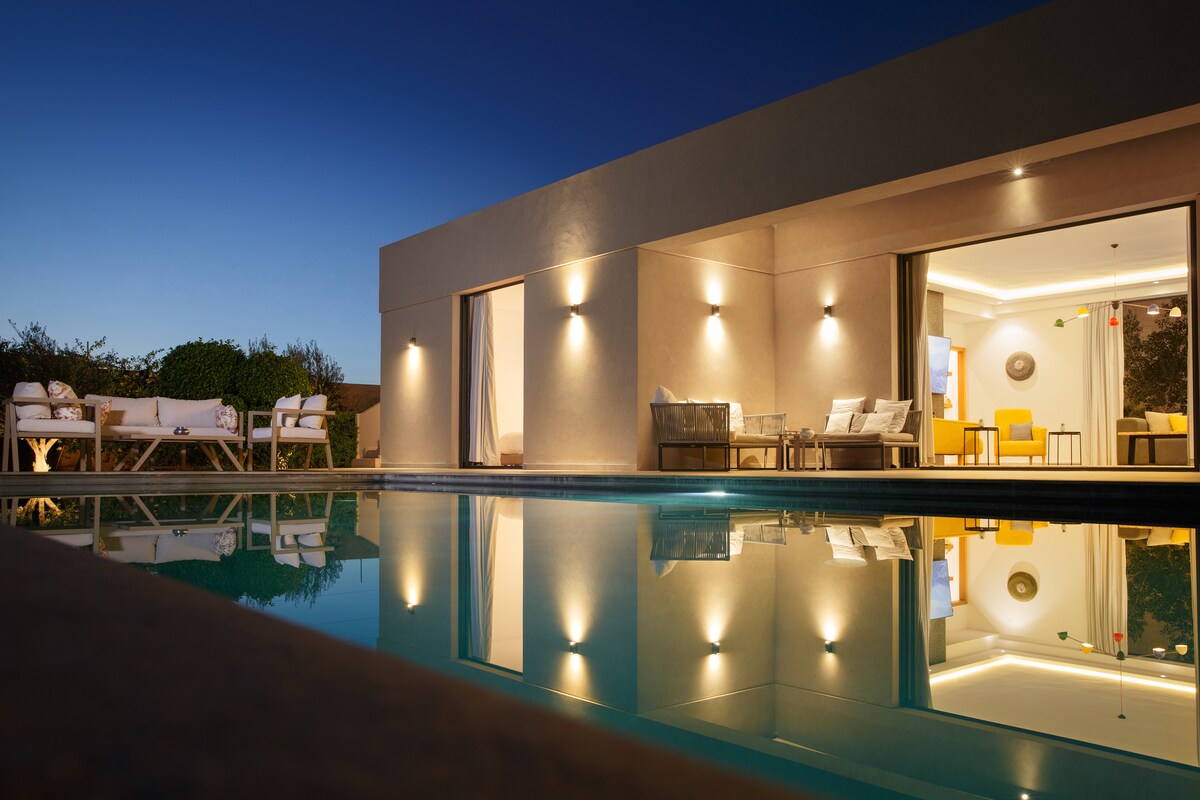 The exterior of the villa is reflected in the calm waters of the heated pool, showcasing a modern design with soft ambient lighting. Brightly colored chairs and a tasteful seating area are visible, promoting a serene outdoor space that invites relaxation.