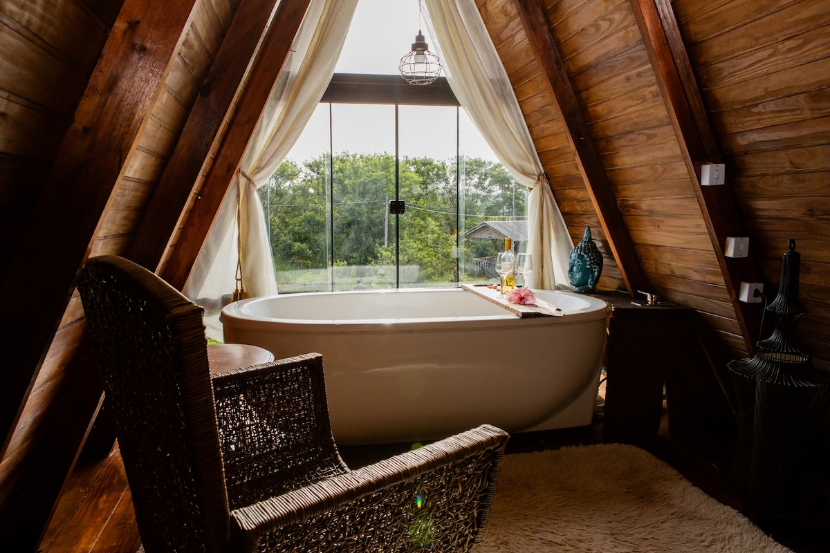 A spacious bathroom is highlighted by an elegant freestanding bathtub positioned near large windows, offering views of the surrounding greenery. A cozy chair complements the space, and soft natural light enhances the warm wooden interior.