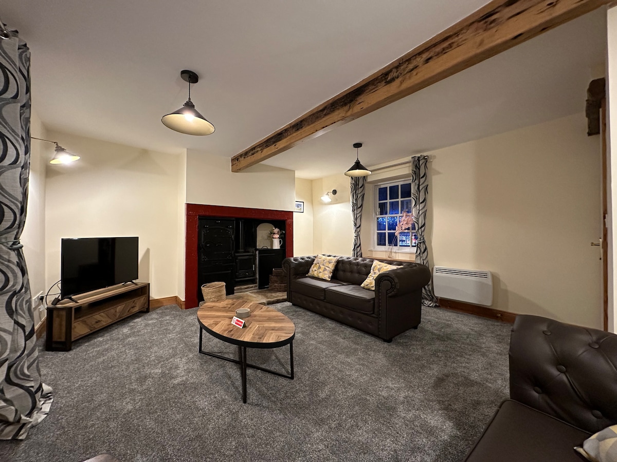 A spacious living room is showcased, featuring a comfortable leather sofa and a wooden coffee table at its center. A large flat-screen TV is positioned against the wall, complemented by an ornate log-burning stove. Windows with patterned drapes allow natural light to illuminate the area.