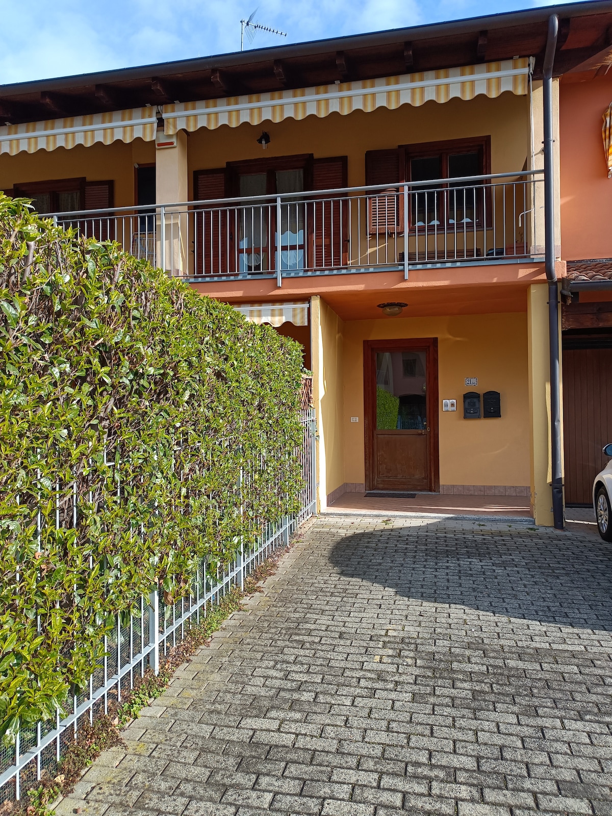 A welcoming entrance is framed by lush green hedges and a paved pathway. The building features a covered porch with a railing, while mailbox units are positioned near the door. The exterior displays a harmonious blend of warm colors and inviting architectural details.