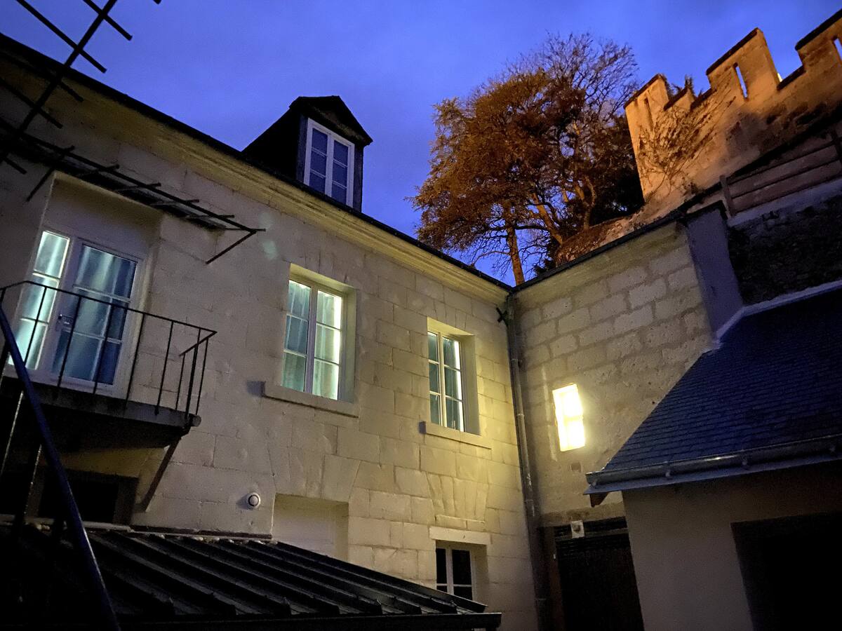 The courtyard of the property is illuminated at dusk, highlighting the stone walls and windows of the building. A tree can be seen above the structure, with the outline of a castle wall in the background, creating a serene atmosphere.