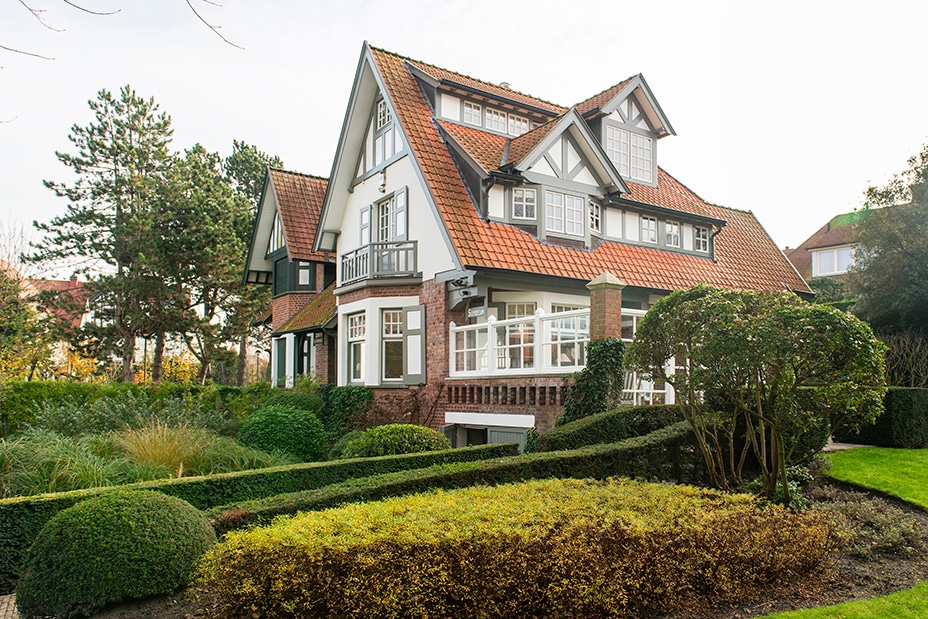 The exterior of a charming two-story villa is presented, featuring a combination of brick and wood elements. A sloping roof is adorned with terracotta tiles, while large windows allow natural light to fill the interior. Lush greenery surrounds the property, enhancing its serene setting.