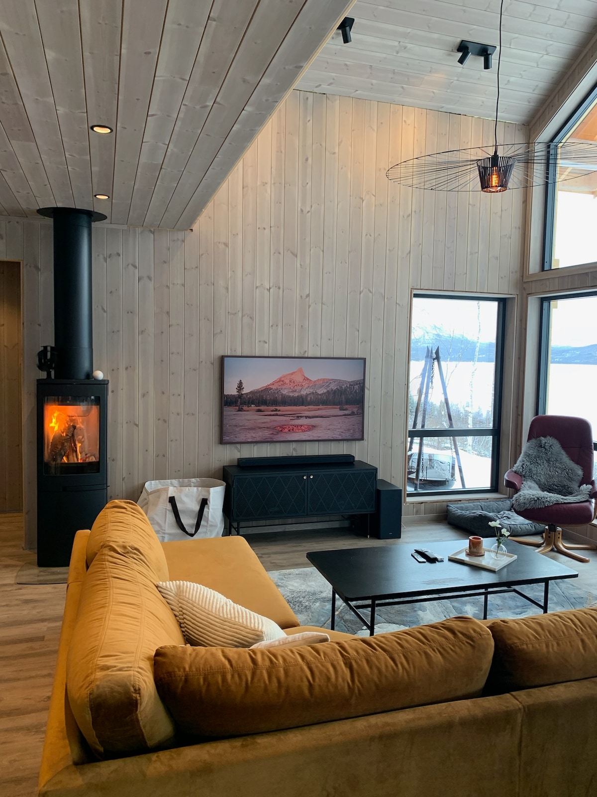 A spacious living room features large windows allowing natural light to fill the area. A modern couch is situated in front of a sleek coffee table. A fireplace adds warmth, while a television is mounted on the wall. A comfortable armchair offers additional seating.