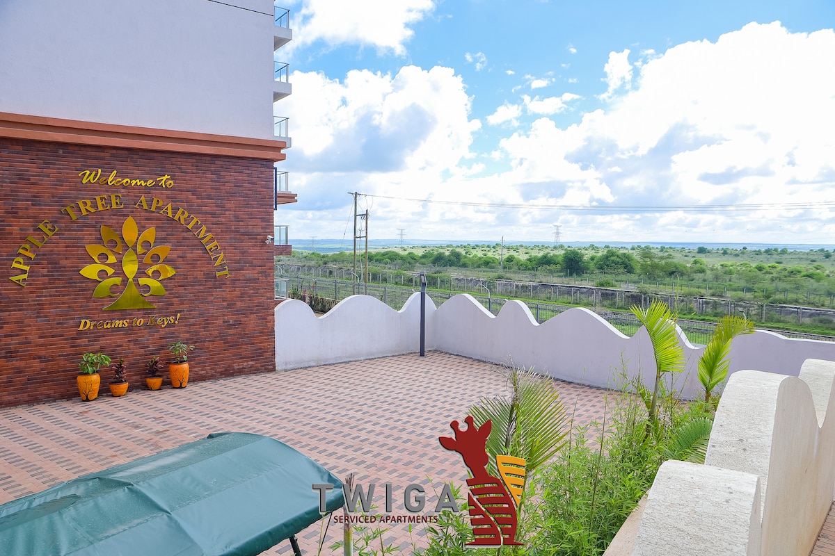 An outdoor terrace is showcased with a patterned tiled floor, surrounded by potted plants. A large sign welcomes guests to Apile Tree Apartment, with a backdrop of expansive greenery under a partly cloudy sky.