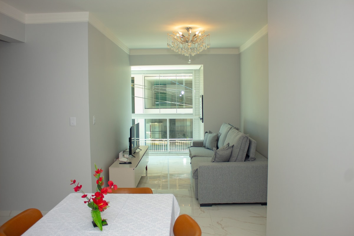 An open living space is visible, featuring a light gray sofa set and a television mounted on the wall. A dining table is set with a floral centerpiece, while large windows provide natural light to the airy environment. Decorative elements add a touch of elegance.