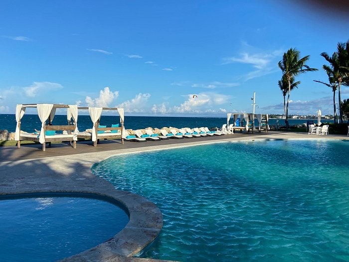 A refreshing beachfront pool is highlighted, surrounded by lounge chairs and shaded cabanas. The clear water reflects the sky, and the ocean is visible in the background, complemented by palm trees swaying gently in the breeze.