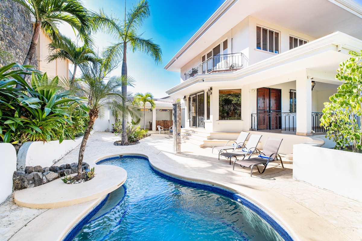 An expansive terrace features a refreshing pool, surrounded by tropical plants and palm trees. Sunbeds are arranged along the pool, creating a serene outdoor space. The villa’s two-story structure stands in the background, with covered patios and large windows inviting natural light.