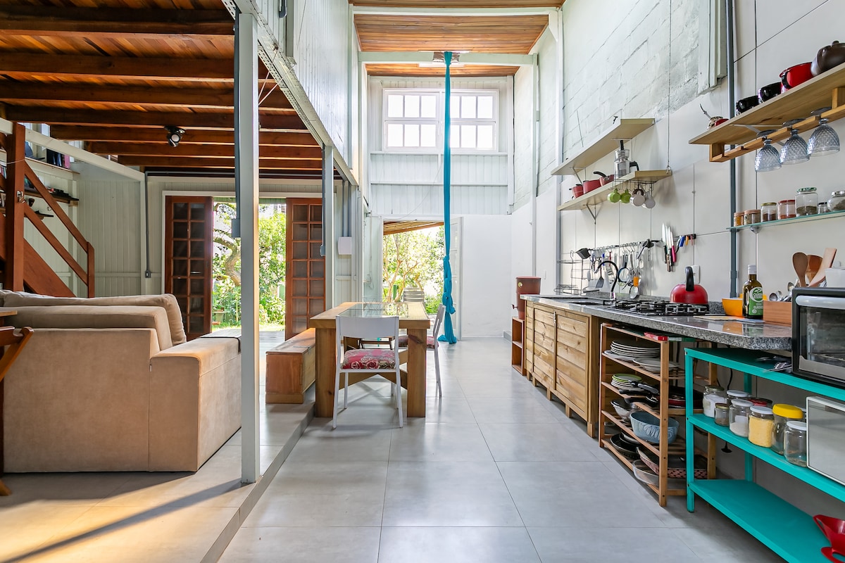 The open-concept living area features a spacious kitchen and dining area, highlighted by natural light streaming through large windows. A wooden table is positioned centrally, surrounded by comfortable seating, while shelves display a variety of kitchenware and decor.