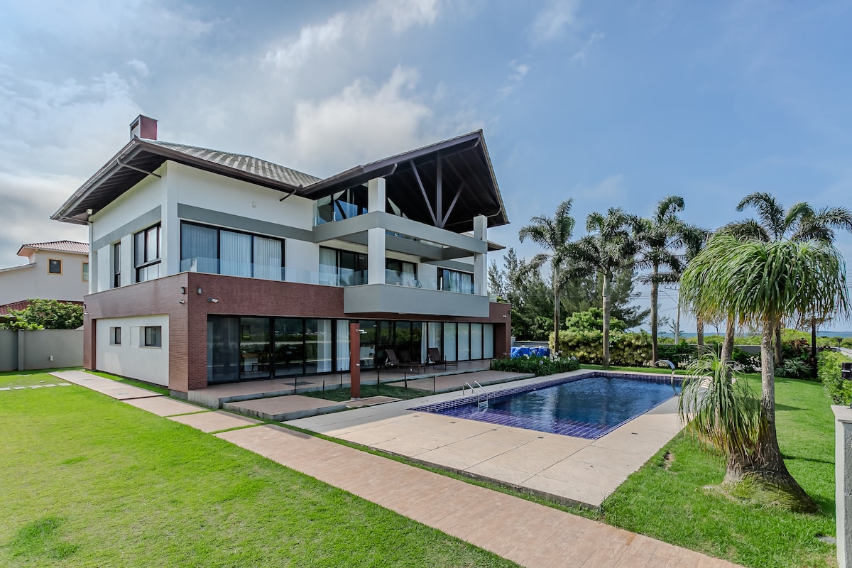 A modern two-story house is presented with large glass windows and a spacious outdoor area. The private pool reflects the sky, surrounded by well-maintained grass and tropical landscaping, enhancing the serene environment of this beachfront property.