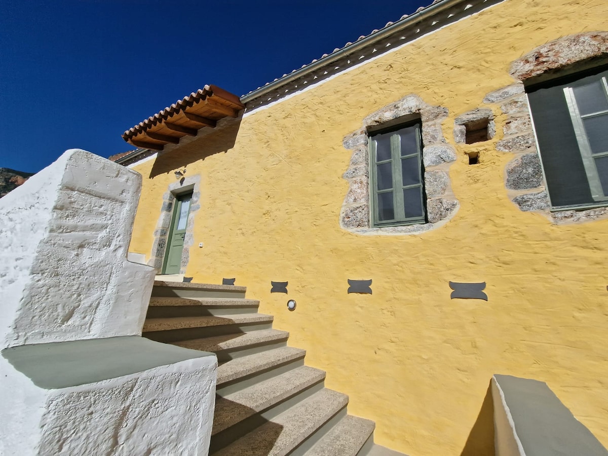 A bright yellow exterior wall is complemented by stone accents and large windows. A staircase leads up towards a door framed by a wooden awning, while unique stonework features add character to the architecture.