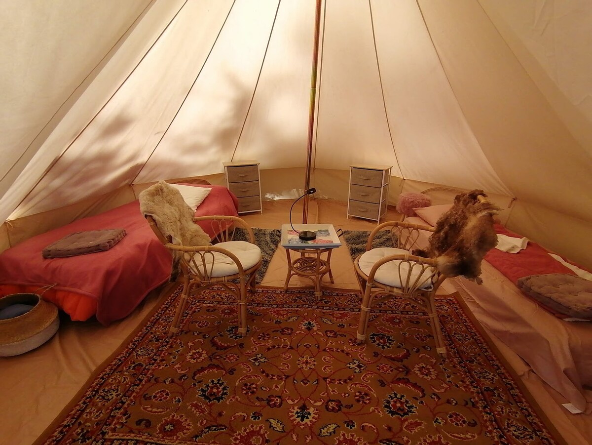 An airy glamping tent features two beds with vibrant bedding and cozy throws. A small table stands between two rattan chairs, complemented by decorative cushions. A woven basket sits on the floor, and a colorful rug adds warmth to the space.