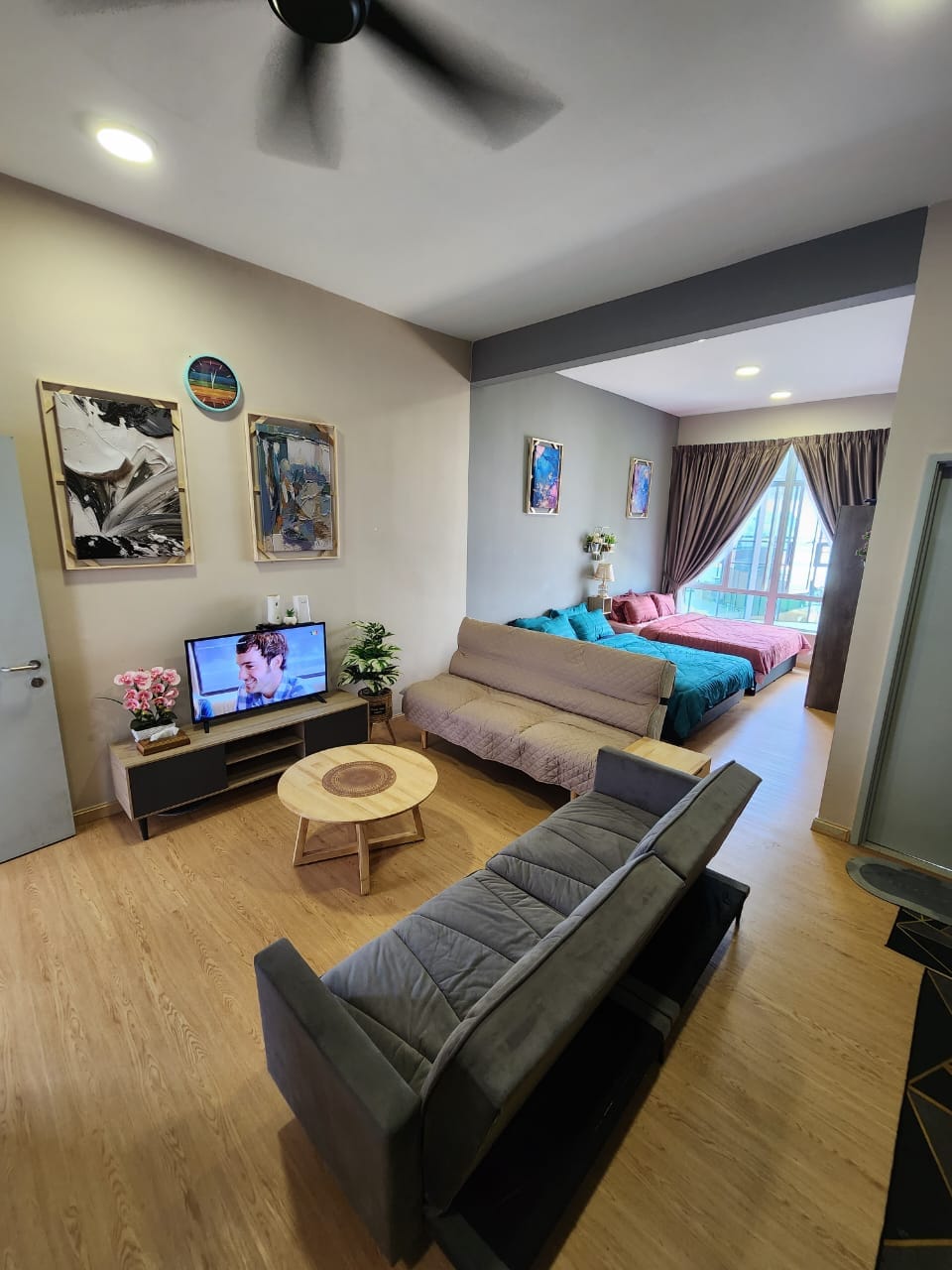 A cozy studio layout features two queen beds dressed in colorful bedding, alongside a spacious sofa and modern television. Large windows allow natural light to fill the room, complemented by tasteful artwork on the walls and a circular table in the center.