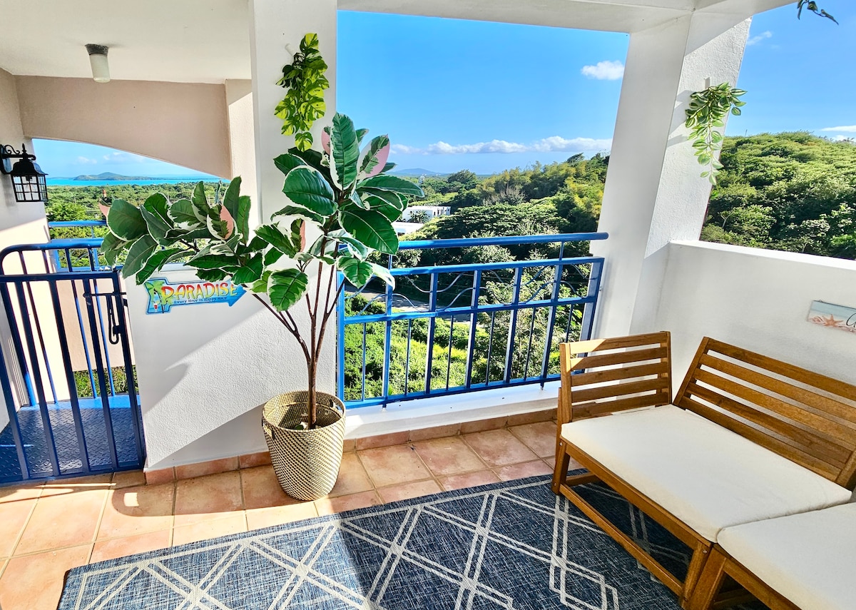 A welcoming outdoor balcony space features a comfortable seating area with a light-colored sofa and decorative plants. Scenic views of lush greenery and distant mountains are framed by a blue railing, allowing plenty of natural light and fresh air to flow through.