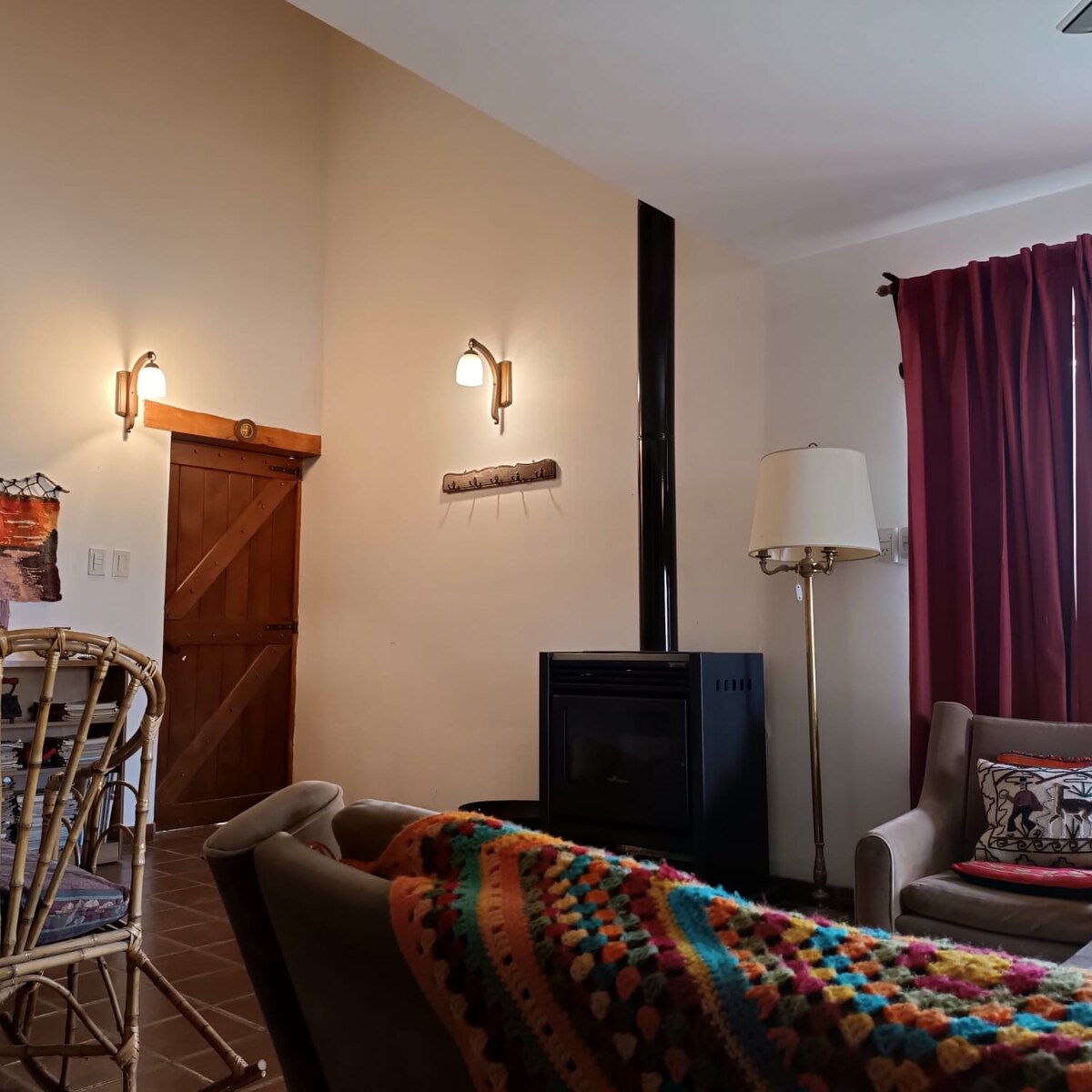 A comfortable living area features a wood-burning stove against a light-toned wall. A plush sofa is adorned with a colorful blanket, complemented by a nearby stylish lamp. Rich red curtains frame the window, adding warmth to the space.