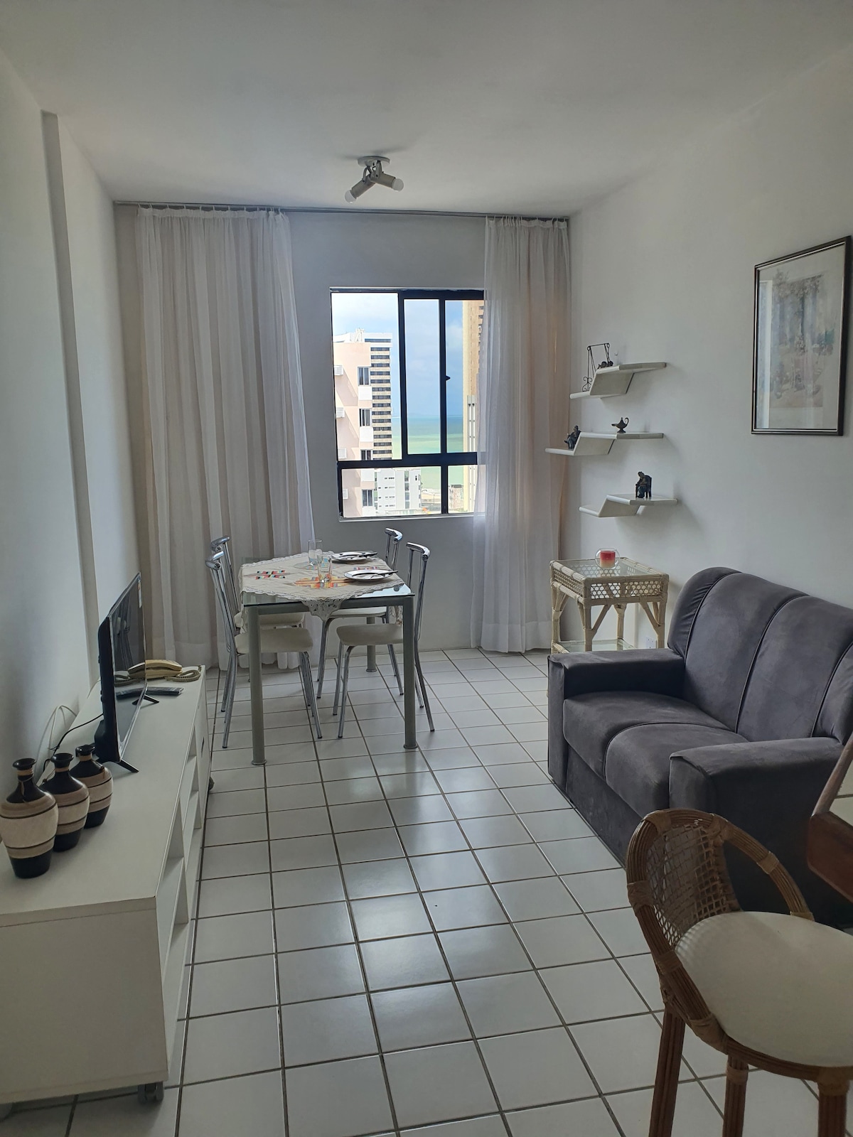 A bright living space features a large window with sheer curtains, offering views of the city. A gray sofa is positioned opposite a white TV stand. A dining table with metal chairs is set nearby, complemented by decorative shelves on the wall.
