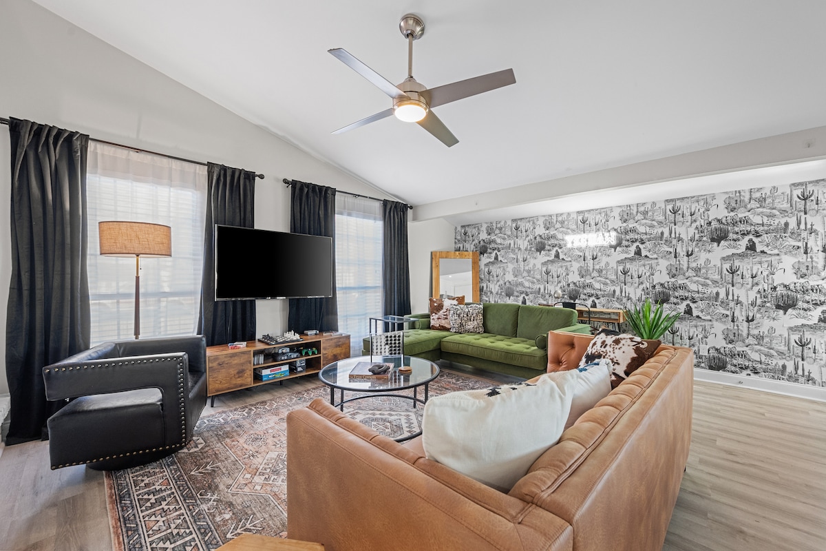 A spacious living area features a mix of comfortable seating, including a leather sofa and an armchair. A large TV is mounted on the wall, and a decorative rug covers the floor. Floor-to-ceiling windows with dark curtains allow natural light to fill the room.