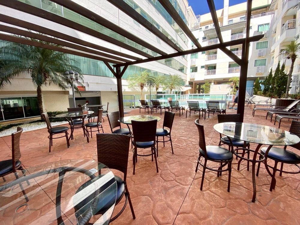 An outdoor seating area is presented, featuring multiple round glass-top tables surrounded by comfortable chairs. A pergola provides partial shade, while tropical plants add greenery to the space. A pool area is visible in the background, enhancing the outdoor ambience.