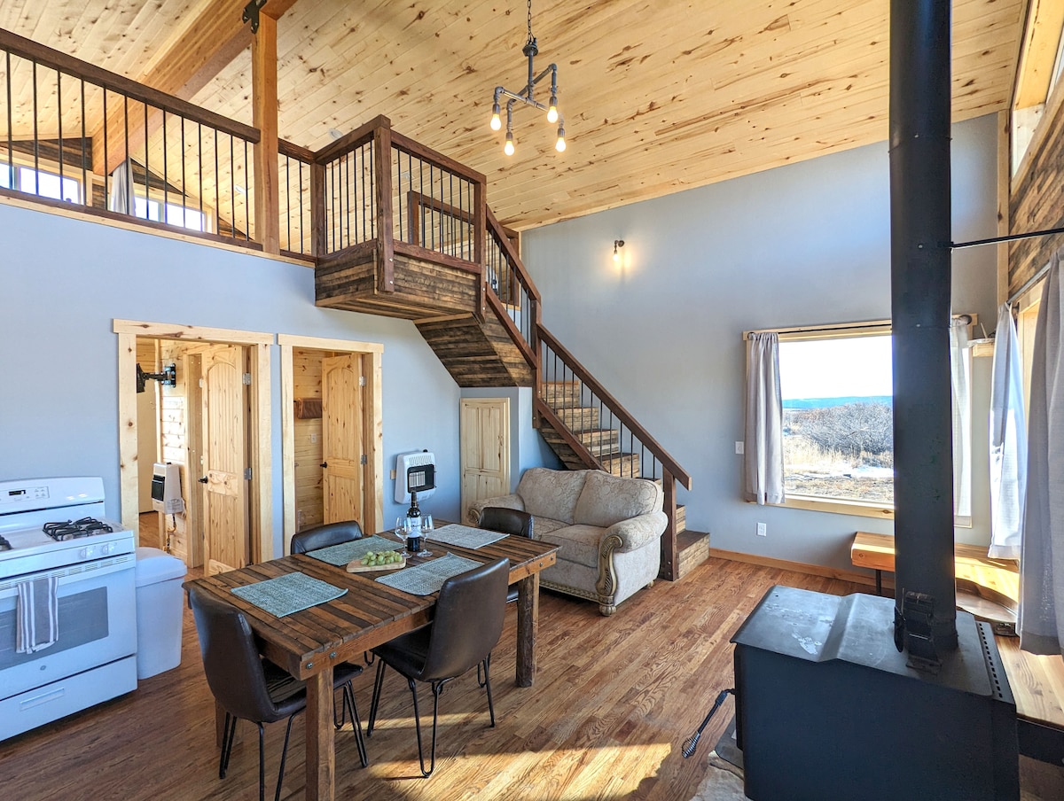 An open living area showcases a rustic dining table surrounded by seating, with a cozy couch nearby. Natural wood beams and a staircase draw the eye upwards, while large windows provide ample light and a view of the serene surroundings.