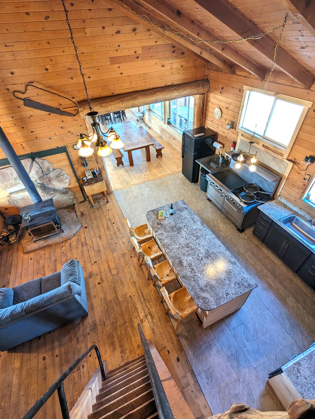 An aerial view of the spacious kitchen and dining area shows wooden floors and walls, complemented by natural lighting from large windows. A kitchen island with seating faces a dining table. A cozy wood-burning stove is positioned nearby, enhancing the functional layout.