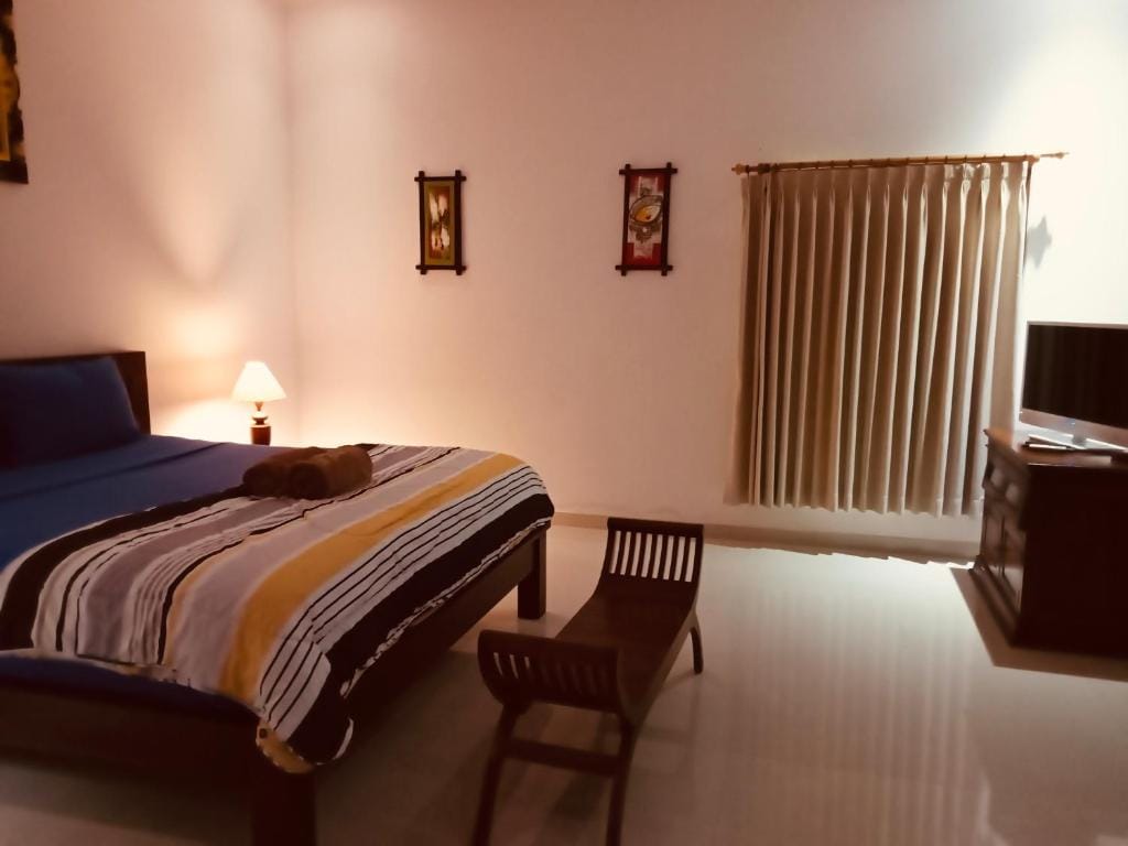 A bright bedroom features a spacious bed with a striped blanket. A versatile chair and a wooden table are present beside the bed. Soft lighting is provided by a bedside lamp, and a television is positioned on a dark wood cabinet. Vertical blinds allow for controlled natural light.