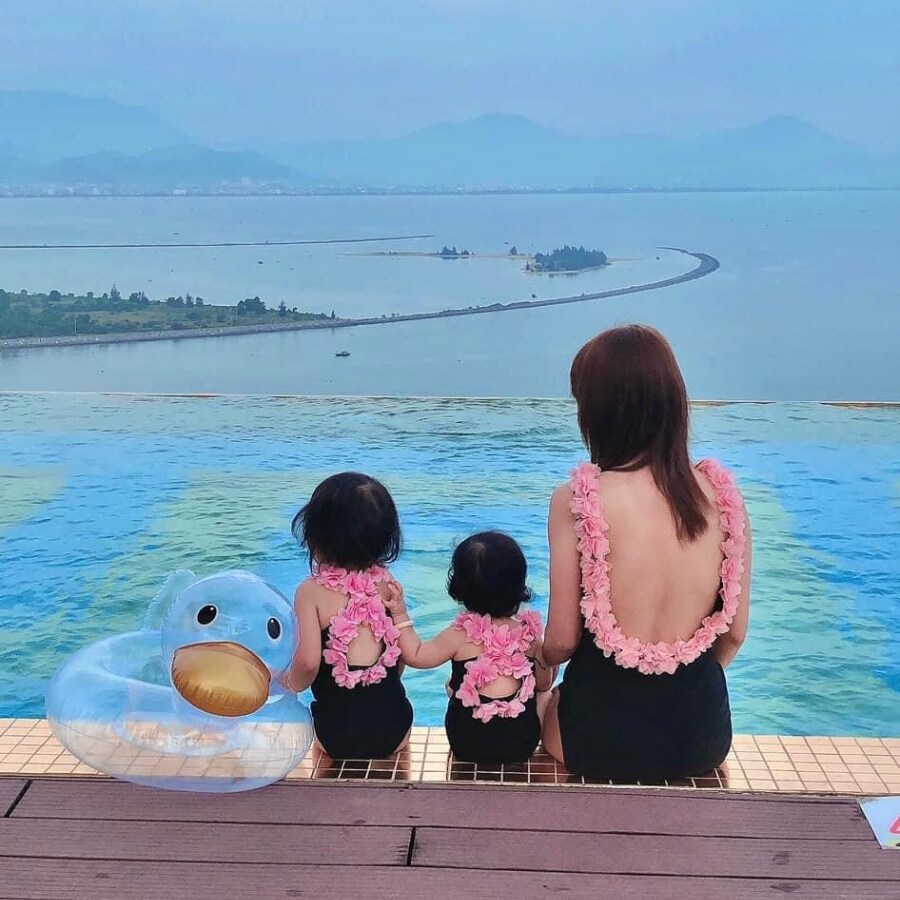 A woman and two children are seated at the edge of an infinity pool, overlooking a scenic view of the water and distant islands. The woman and children are in matching swimwear adorned with floral accents. A blue inflatable pool float in the shape of a duck is placed nearby.