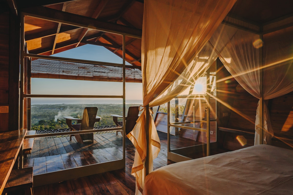 A spacious cabin interior reveals a stunning view through large glass doors, allowing natural light to illuminate the room. The wooden beams of the ceiling provide a warm touch, while sheer curtains gently frame the view of the expansive landscape beyond.