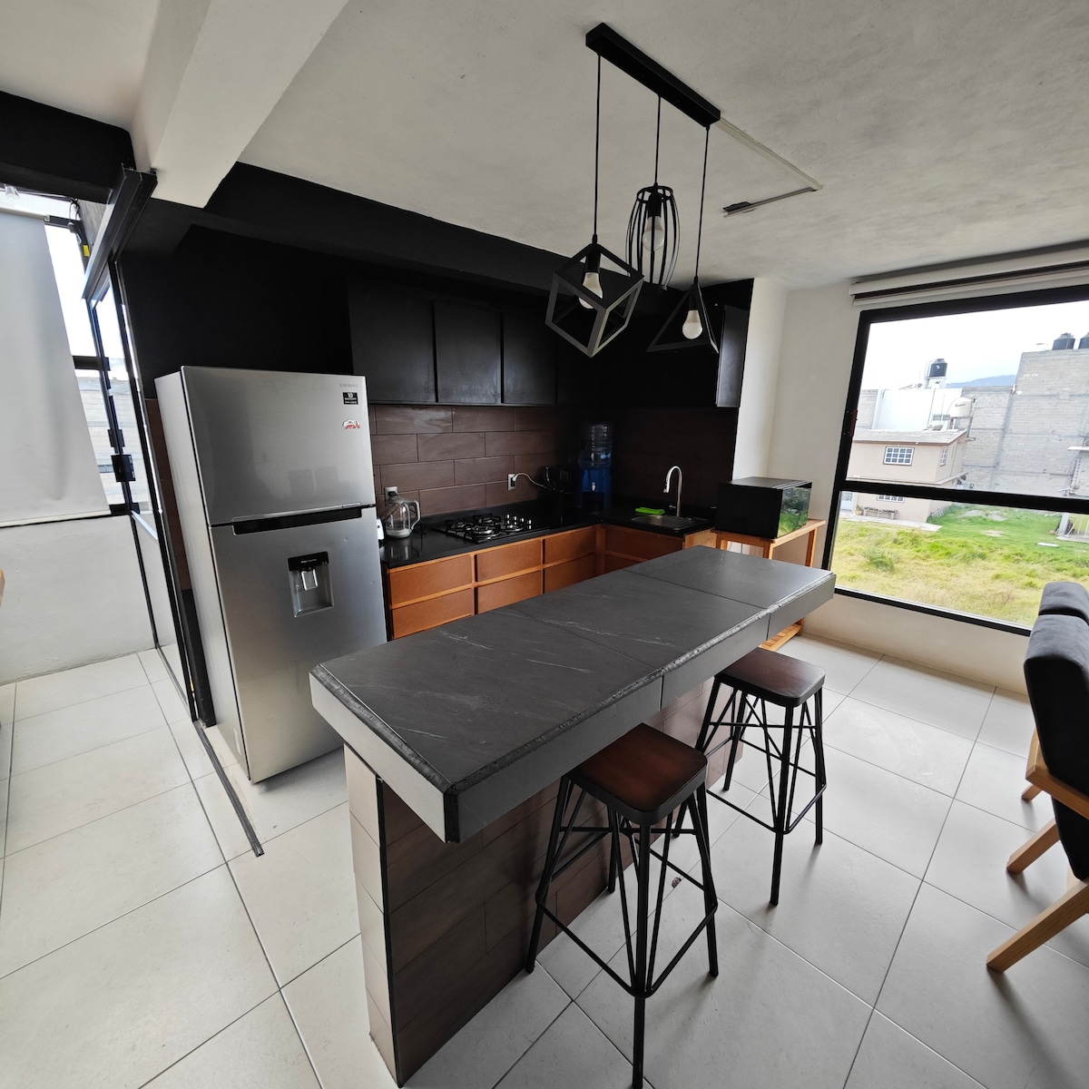 An open kitchen area features modern cabinetry and a sleek countertop. A stainless steel refrigerator and a stove are included. Two wooden bar stools are positioned at the counter, providing a casual dining option. Large windows allow natural light to brighten the space.