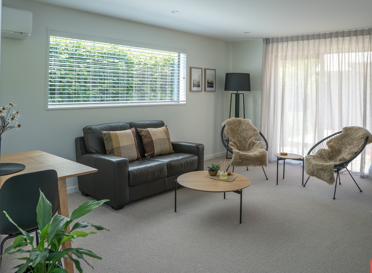 A spacious living room is furnished with a dark leather sofa and two accent chairs featuring soft throws. A light wood coffee table is centered on a neutral-toned carpet. Natural light enters through large windows covered with sheer curtains, creating an inviting atmosphere.