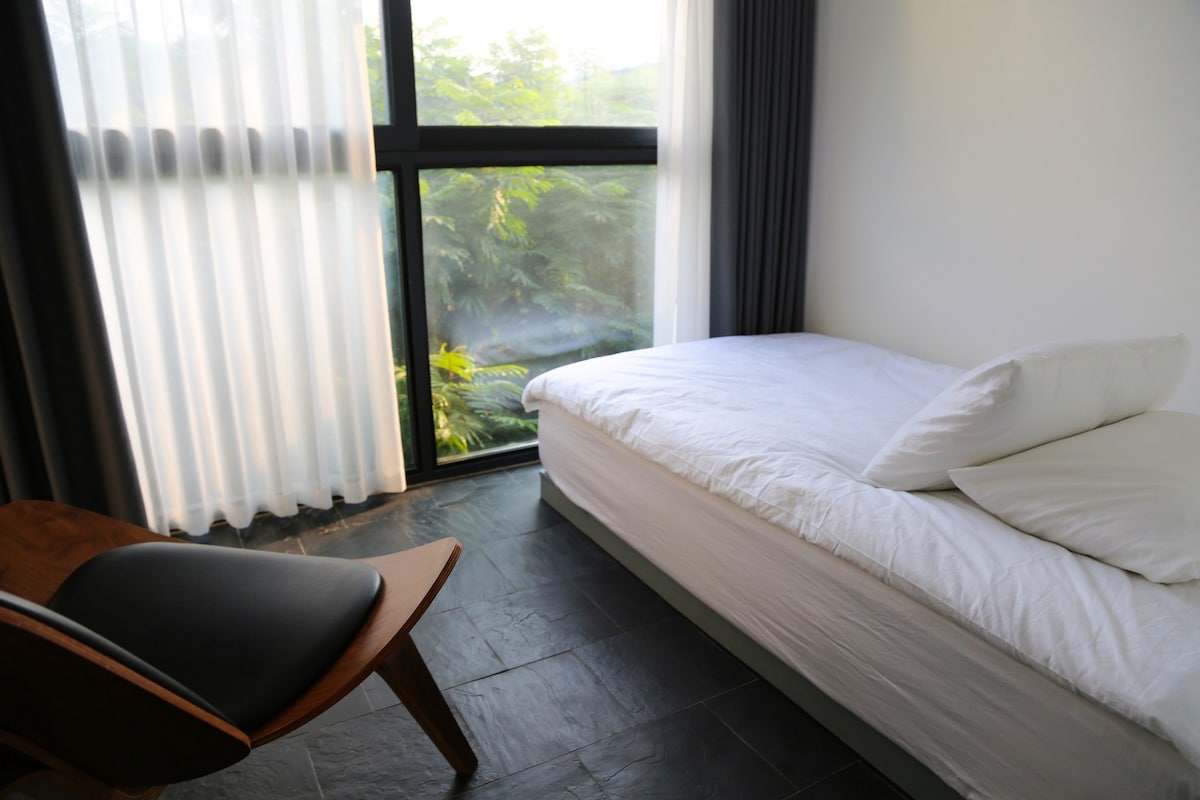 A minimalist bedroom features a spacious bed with crisp white linens positioned beside a large window. Soft natural light filters through sheer curtains, illuminating the room. A wooden chair is placed at an angle, adding a touch of elegance to the contemporary design.