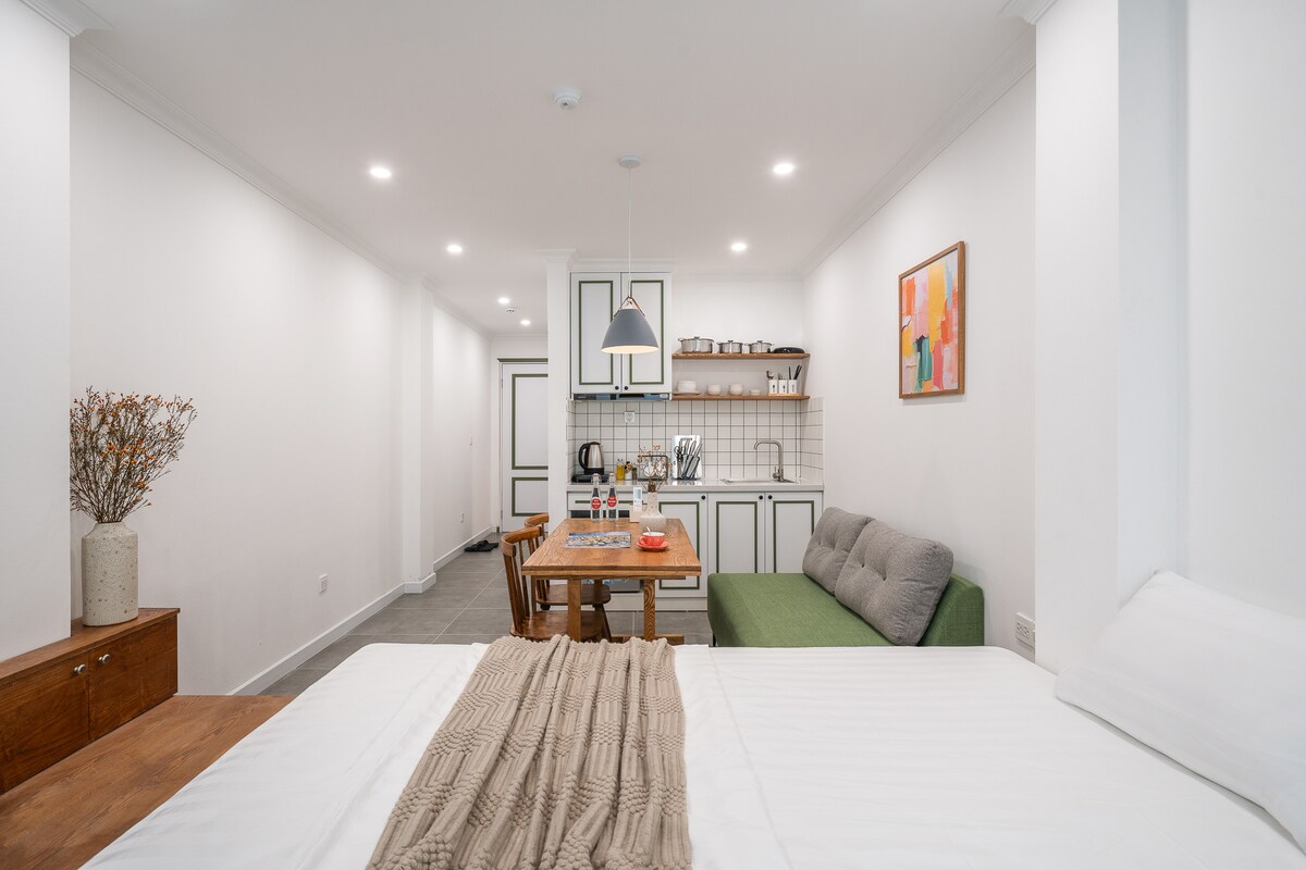 An open-concept living space features a light-colored queen-size bed with a textured throw. A green sofa sits nearby, accompanied by a wooden dining table set for four. A neatly arranged kitchen area is visible in the background, along with decorative artwork on the wall.