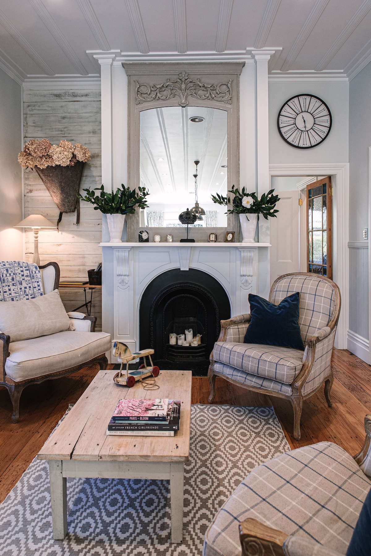 A charming lounge area features two upholstered chairs with plaid patterns, positioned around a wooden coffee table. A classic fireplace, framed by a decorative mirror, serves as the focal point, complemented by fresh flowers in elegant vases. A patterned rug adds warmth to the space.