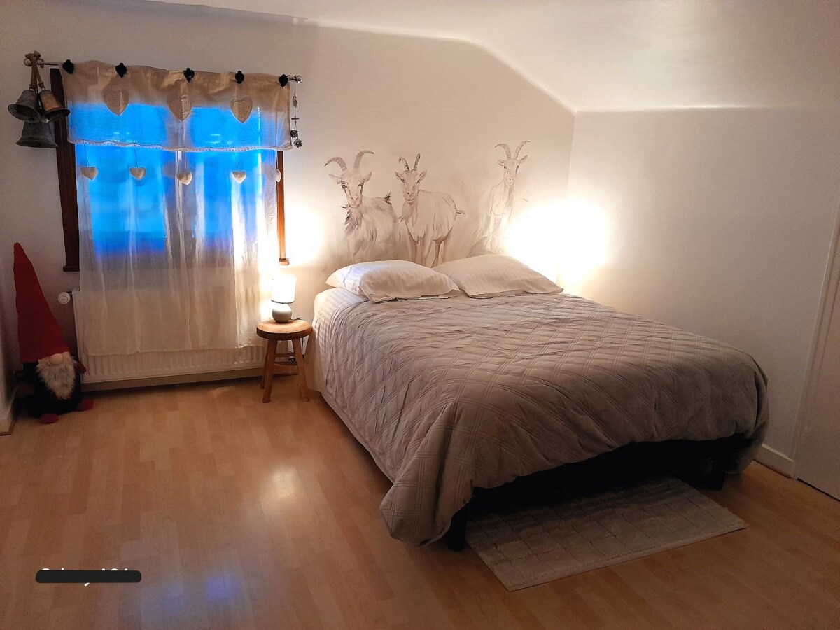 A cozy bedroom features a spacious bed covered with a light quilt and a decorative wall showcasing goat motifs. Natural light enters through a curtained window, while a bedside lamp provides soft illumination. Light wooden flooring enhances the room's serene atmosphere.