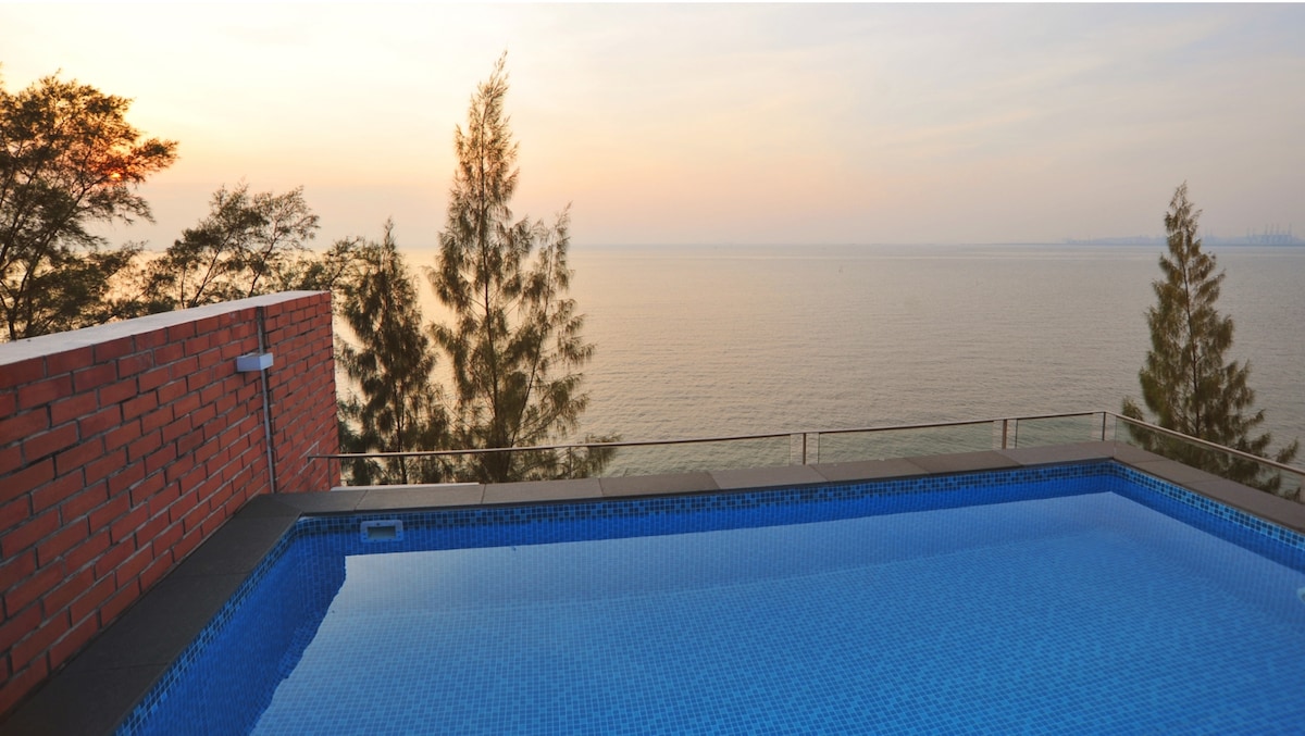 The rooftop pool is framed by brick walls, offering inviting blue water reflecting the soft colors of the sunset. Views of the serene sea and silhouetted trees create a peaceful atmosphere, enhancing the space's natural beauty.