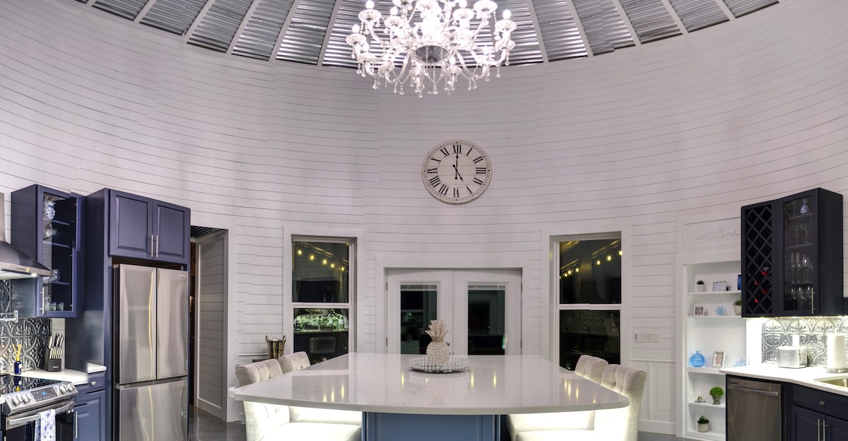 A modern kitchen features a spacious island at its center, surrounded by soft seating. A sophisticated chandelier hangs from a curved, metallic ceiling, complemented by a large clock on the wall. Bright cabinetry and warm lighting enhance the inviting atmosphere of this unique space.