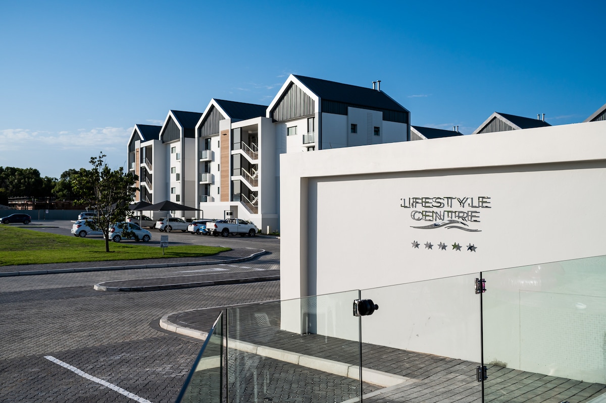 The Lifestyle Centre is presented with modern architectural designs, featuring multiple levels of apartments adorned with gabled roofs. A prominent sign identifies the centre, while a spacious parking area accommodates vehicles. Clear blue skies create an inviting backdrop for the contemporary structure.