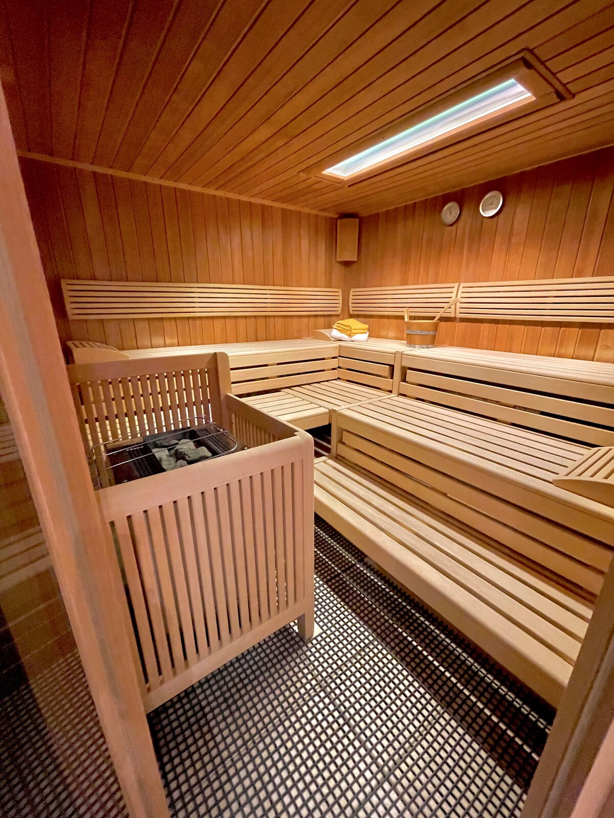 A wooden sauna is shown, featuring several built-in benches with a slatted design. The interior is illuminated by overhead lighting. On one of the benches, towels are neatly arranged, and two clocks are mounted on the walls for timekeeping.