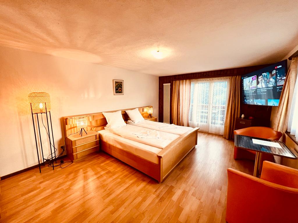 A spacious room features a wooden floor, with a large bed consisting of two separate mattresses. A flat-screen television is mounted on the wall, while a cozy seating area includes orange chairs and a small table. Soft light is provided by a stylish lamp.