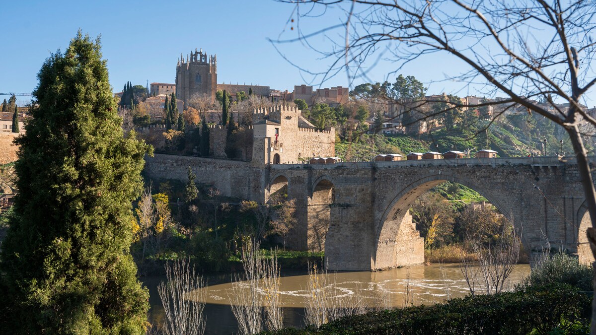 A historic stone bridge spans a calm river, leading towards a hillside adorned with ancient structures and lush greenery. The picturesque landscape features a castle-like building in the distance, while vibrant trees frame the scene, enhancing the natural beauty of Toledo.