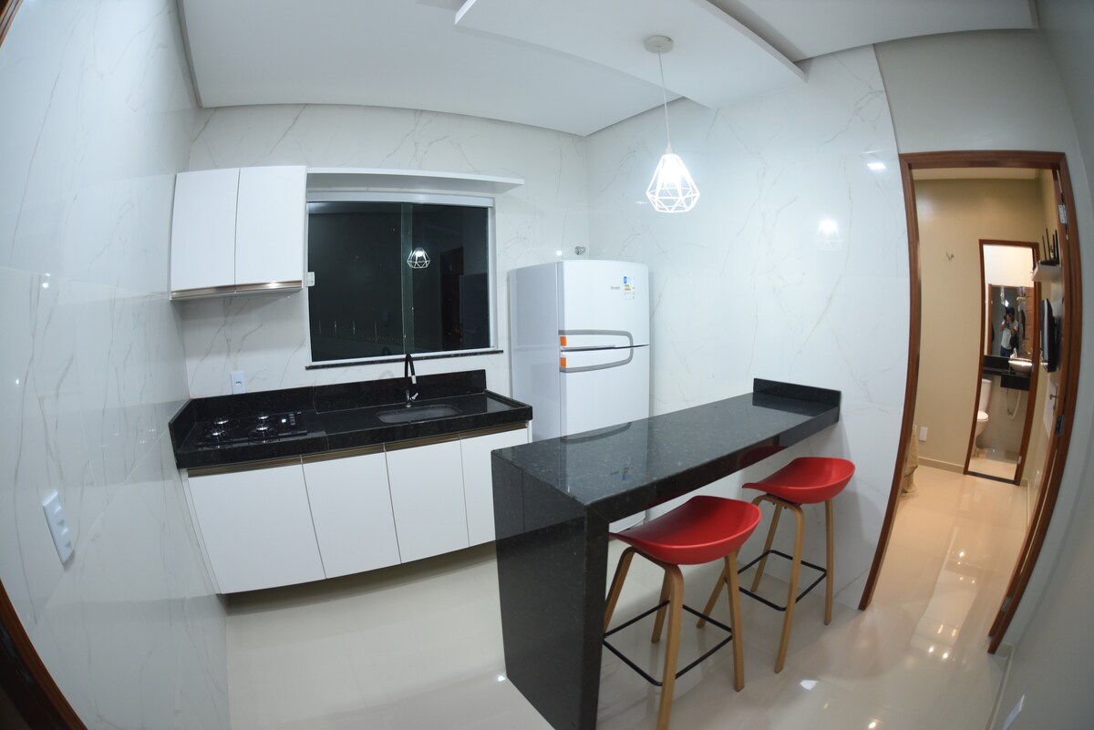 A modern kitchen area features sleek white cabinetry and a black countertop. A large window allows natural light to brighten the space. Two red stools are situated at a comfortable breakfast bar, adding a pop of color to the minimalist design. A doorway leads to the adjacent room.