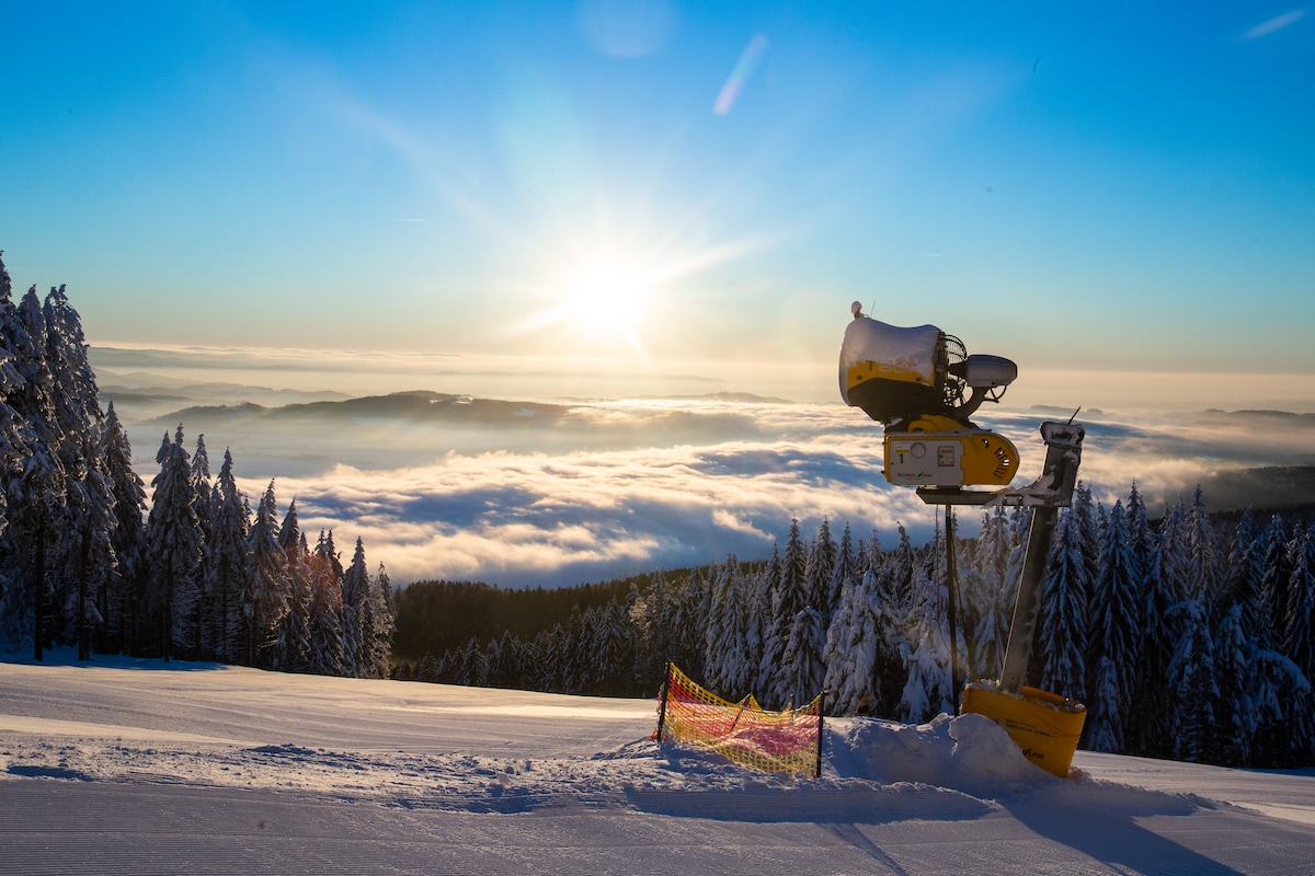 A ski resort setting is captured with a snow-covered slope illuminated by the rising sun. In the foreground, a snow-making machine stands next to a safety barrier, while a blanket of clouds covers the valley below, surrounded by snow-dusted trees.