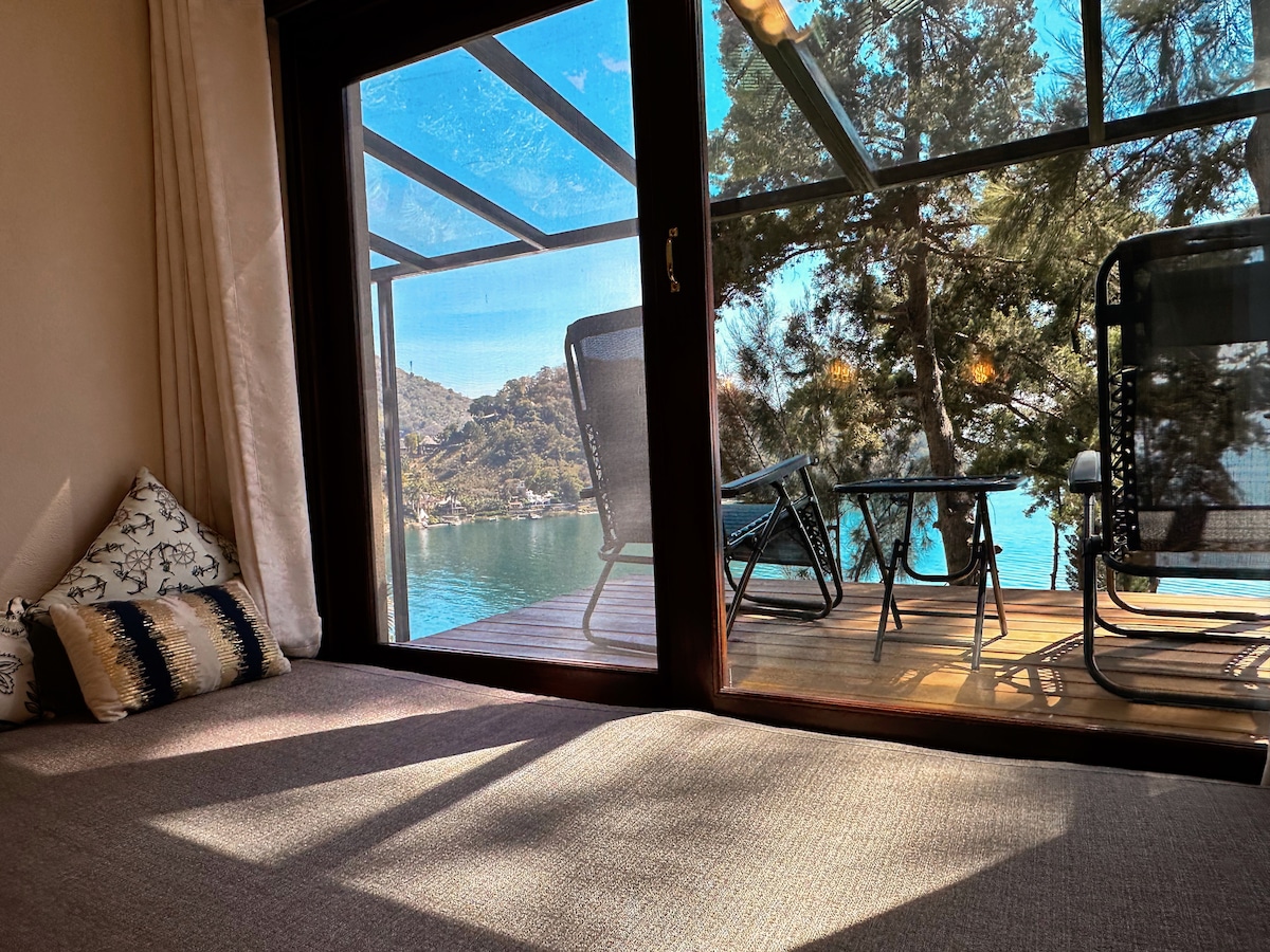A cozy interior space features floor-to-ceiling sliding glass doors, providing a clear view of a private balcony overlooking the lake. Comfortable outdoor seating is visible on the balcony, surrounded by greenery, allowing for a serene connection with nature.