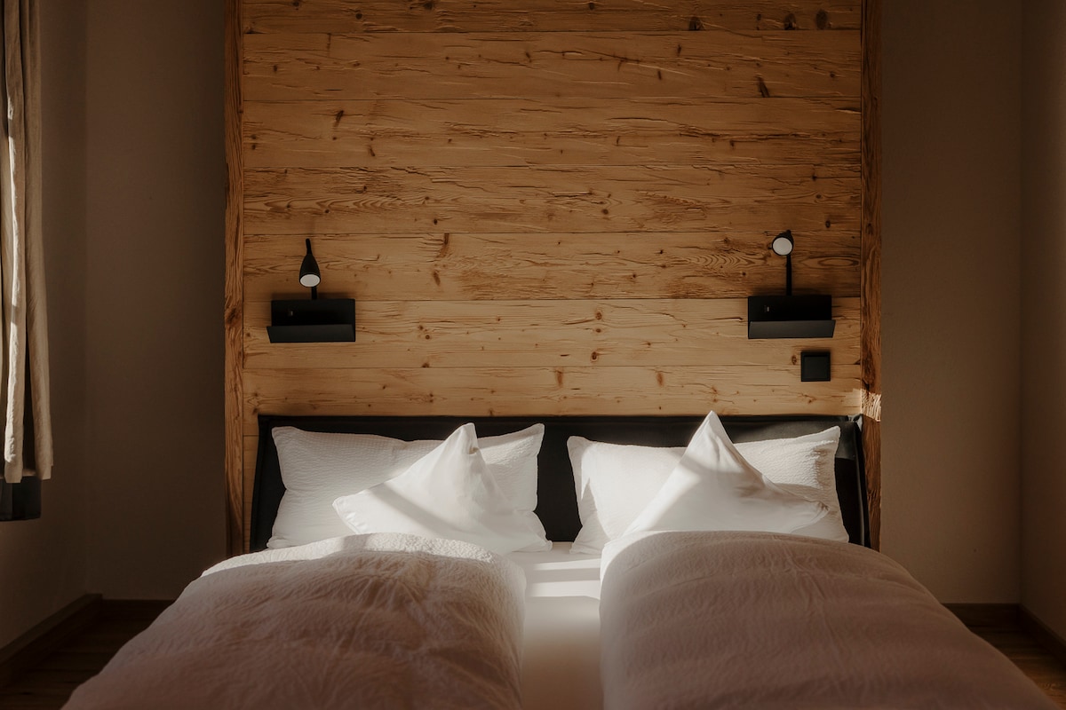 A sleeping area features a wooden accent wall and a spacious bed with white bedding and two pillows. Two minimalist wall lamps provide soft lighting, creating a calming and serene environment. Natural light filters in through a nearby window, highlighting the inviting setting.